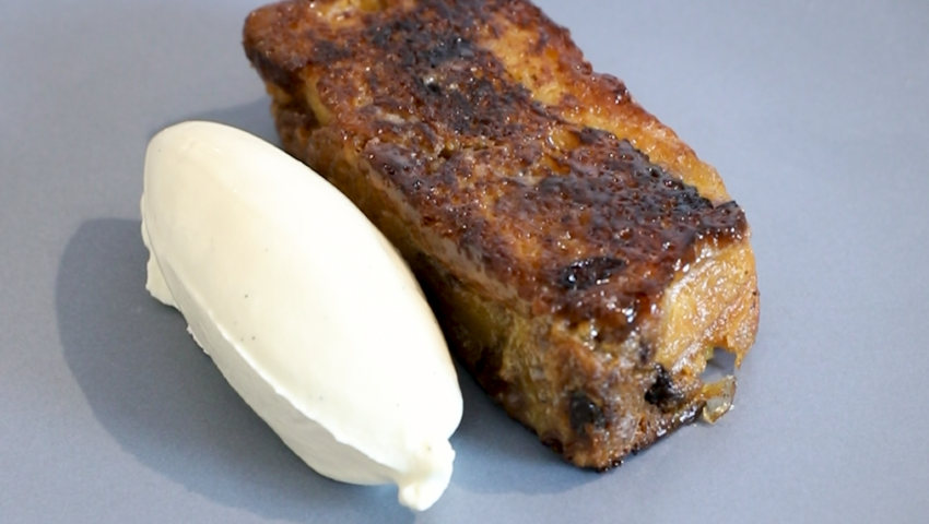An image of a pan fried panettone pudding, with golden brown slices of the dessert arranged on a plate. The pudding has a creamy, layered appearance, with bits of chopped fruit visible.